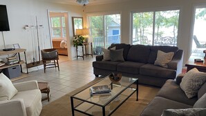 A living room with a sofa, armchairs, a coffee table, and a TV. There is a ceiling fan, a plant, and a window with a view of the outside.
