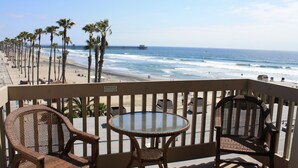 A balcony with wicker chairs and a glass table overlooking a beach with palm trees and a pier.