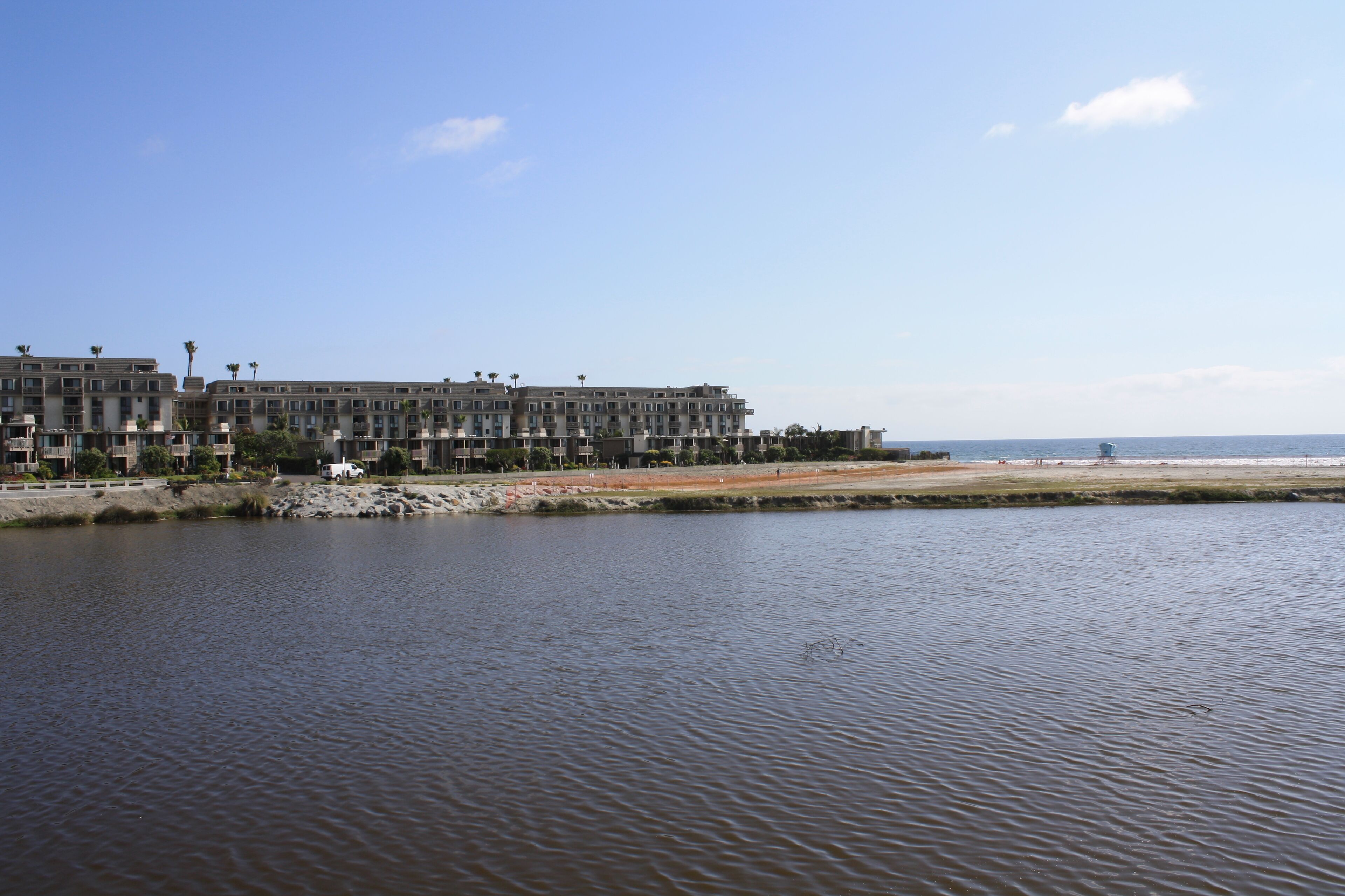 A waterfront hotel with multiple floors, a beach in the background, and a calm water body in the foreground.