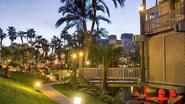 A pathway leading to a building with a wooden deck and a pool area, surrounded by palm trees and well-lit by ambient lighting.