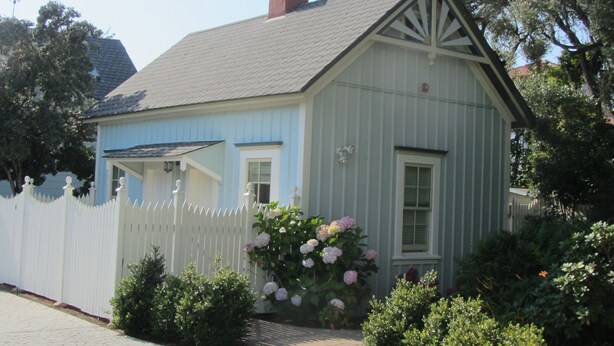 Une maison en bois d’un bleu clair, avec une clôture blanche en piquets, un petit porche et des arbustes d’hortensias roses devant.