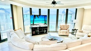 Una sala moderna con un televisor de pantalla plana grande, un sofá seccional, un sillón estampado y una pequeña mesa de centro.