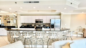 Una cocina moderna con armarios blancos, una isla central con taburetes y un área de comedor con sofá y sillas blancos.