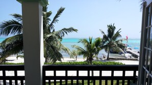 Un balcón con banca que da a una playa con palmeras y un muelle con barcos.