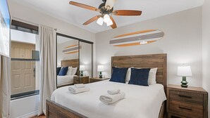 Un dormitorio con cama de madera, dos mesitas de noche con lámparas, un ventilador de techo, un espejo grande y vistas al exterior.