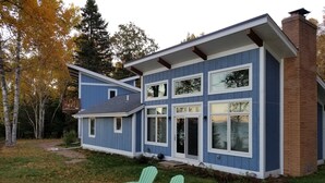 Una casa moderna de dos pisos con fachada azul y detalles blancos, con ventanas amplias y una chimenea de ladrillo.