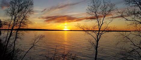 Auringonlasku tyynen vesistön yllä, ja paljaat puut kehystävät maisemaa.