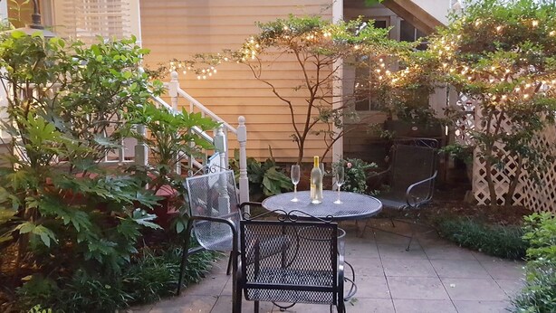 Une terrasse avec une table ronde et des chaises, entourée de verdure et illuminée de guirlandes lumineuses.