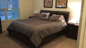 A bedroom with a large bed, a nightstand with a lamp, two framed abstract paintings on the wall, and a window with blinds. - Nice Family Friendly, WalMart and HEB & Wallgreens Walking, Iron Man. (The Woodlands)