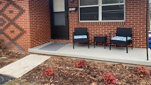 A house with a brick exterior, a black door, and a number 4926 sign above the door. There are two wicker chairs with blue cushions on the porch.