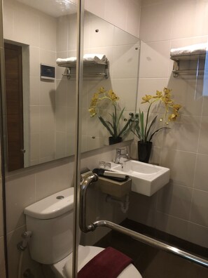 Un baño moderno con cabina de ducha de vidrio, un inodoro blanco, un lavamanos con espejo y orquídeas amarillas sobre el lavamanos.