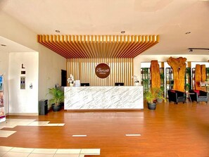 Zona de recepción de un hotel con mostrador de mármol, listones de madera en el techo y plantas en macetas.