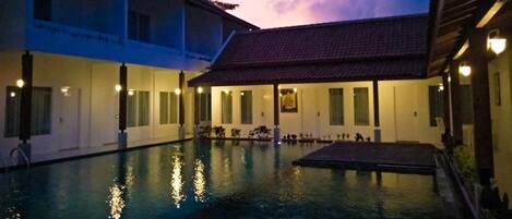A courtyard with a swimming pool, reflecting lights, and a building with a tiled roof.