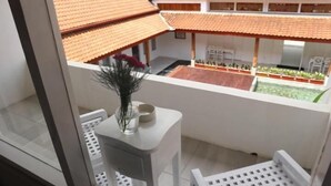 Un petit balcon avec une table blanche, un vase de fleurs et une vue sur une cour intérieure dotée d’un toit en tuiles et de plantes.