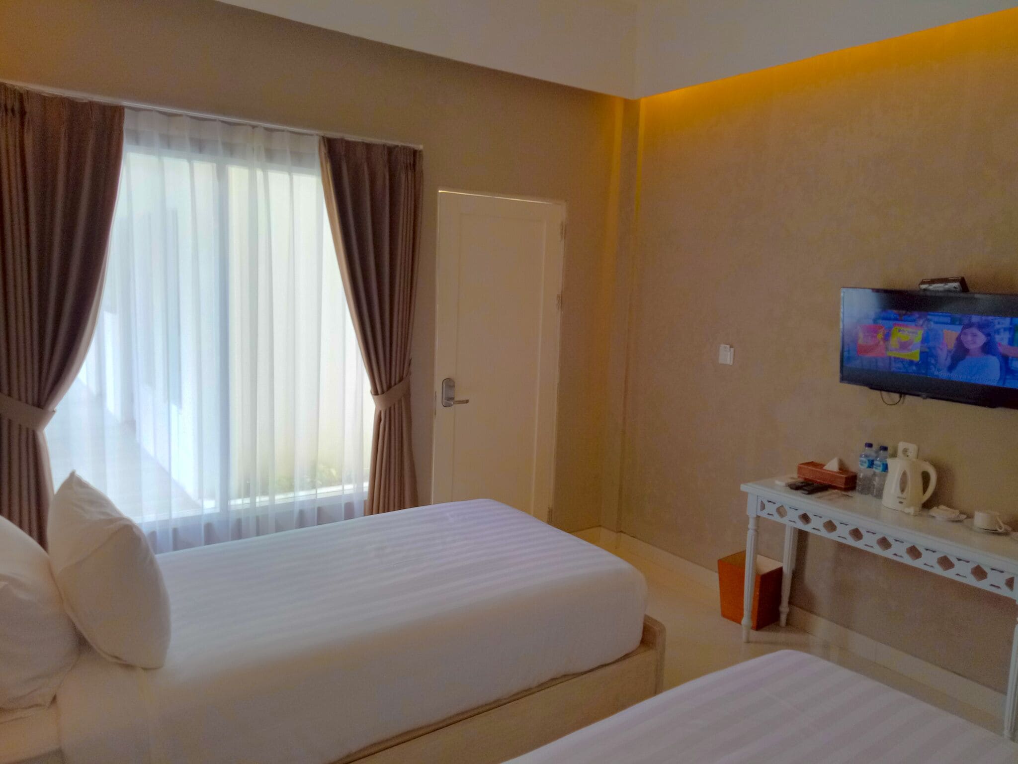 A hotel room with two beds, a TV, a small table with toiletries, and a window with curtains.