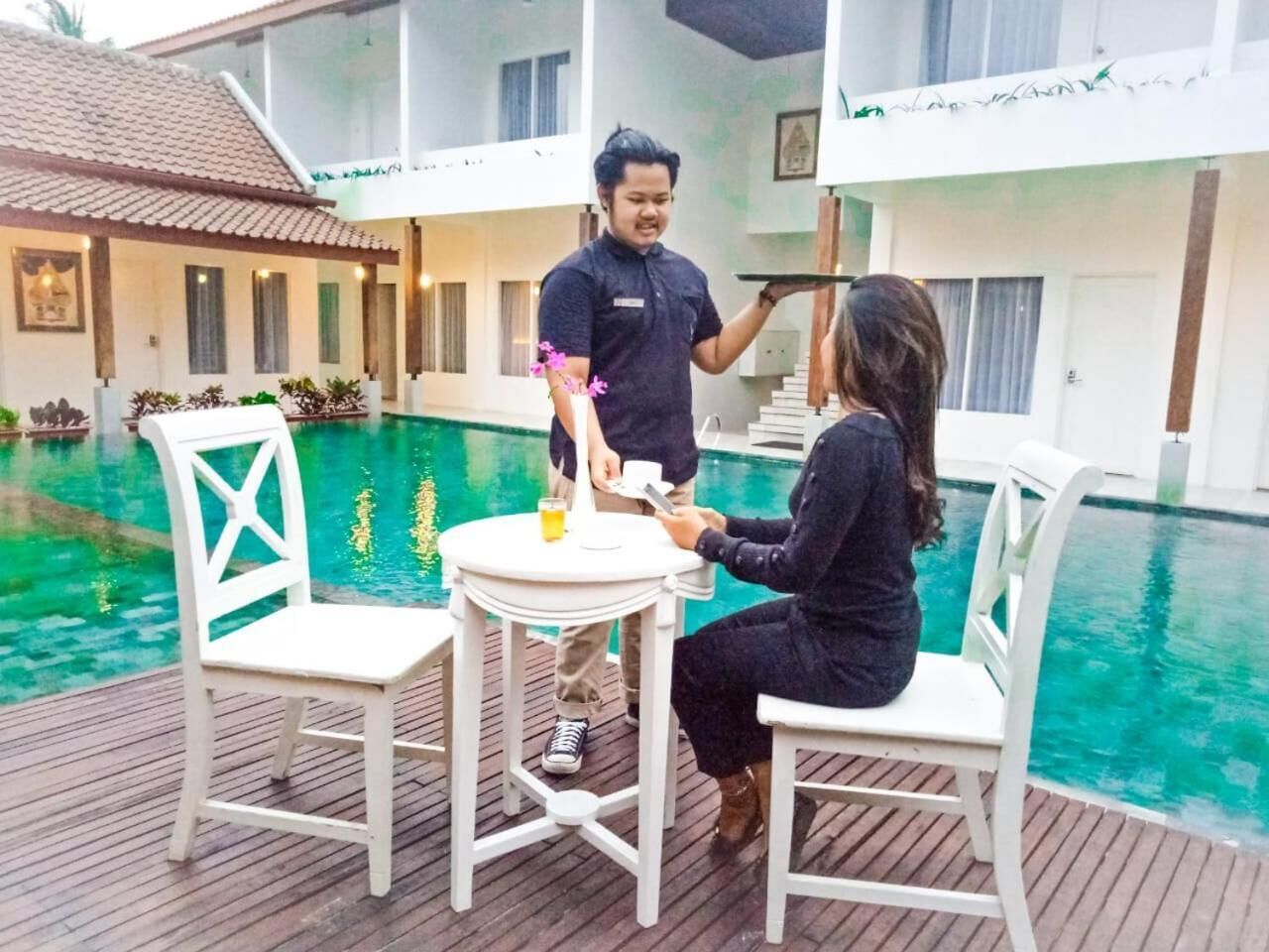 A poolside area with a table and two chairs, a person serving drinks, and a building in the background.