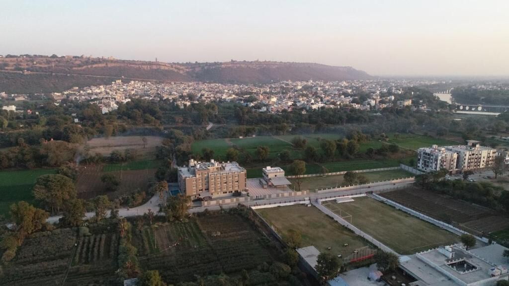 An aerial view of a residential area with a large building complex, green fields, and a distant hill.