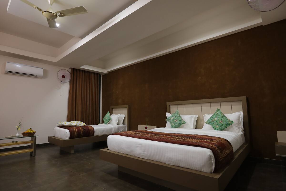A hotel room with two beds, a ceiling fan, an air conditioning unit, and a small table with a vase and a basket.