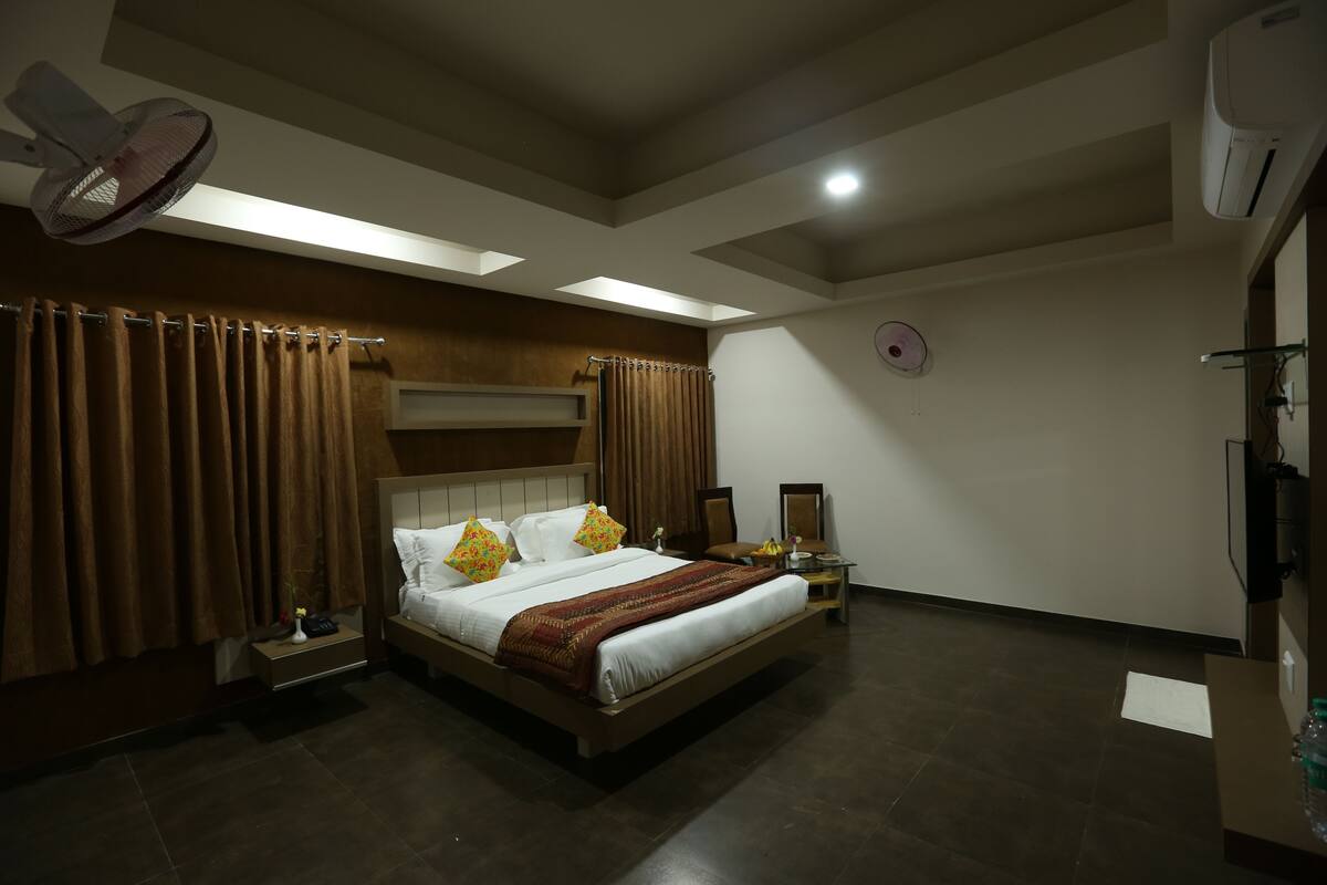 A bedroom with a bed, curtains, a bedside table, a nightstand, a fan, and an air conditioning unit.