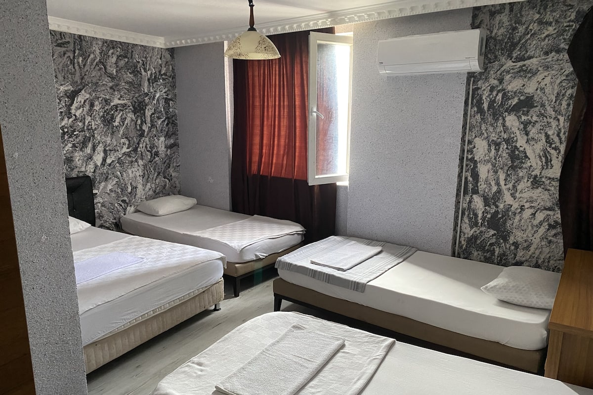 Habitación de hotel con tres camas individuales, una ventana con cortinas rojas, un equipo de aire acondicionado y una pared con diseño de mármol.