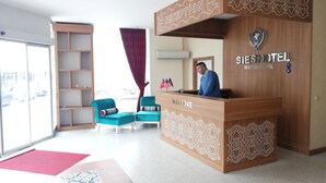 A reception desk with a wooden design and a "Welcome" sign, a blue chair with a striped pillow, and a view of the city through large windows. - Sies Hotel (Antalya)