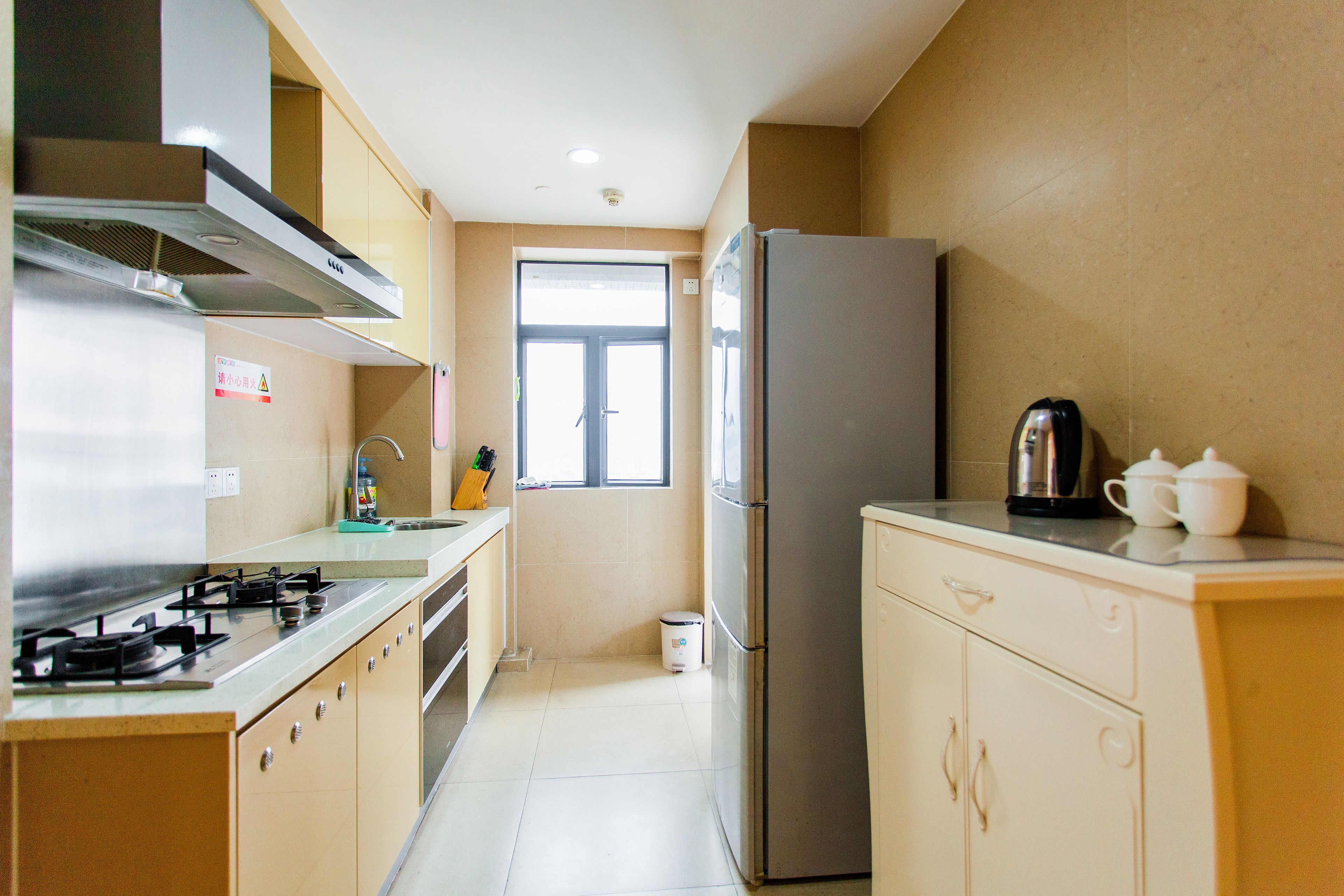 A compact kitchen with a gas stove, stainless steel range hood, a refrigerator, and a window providing natural light.