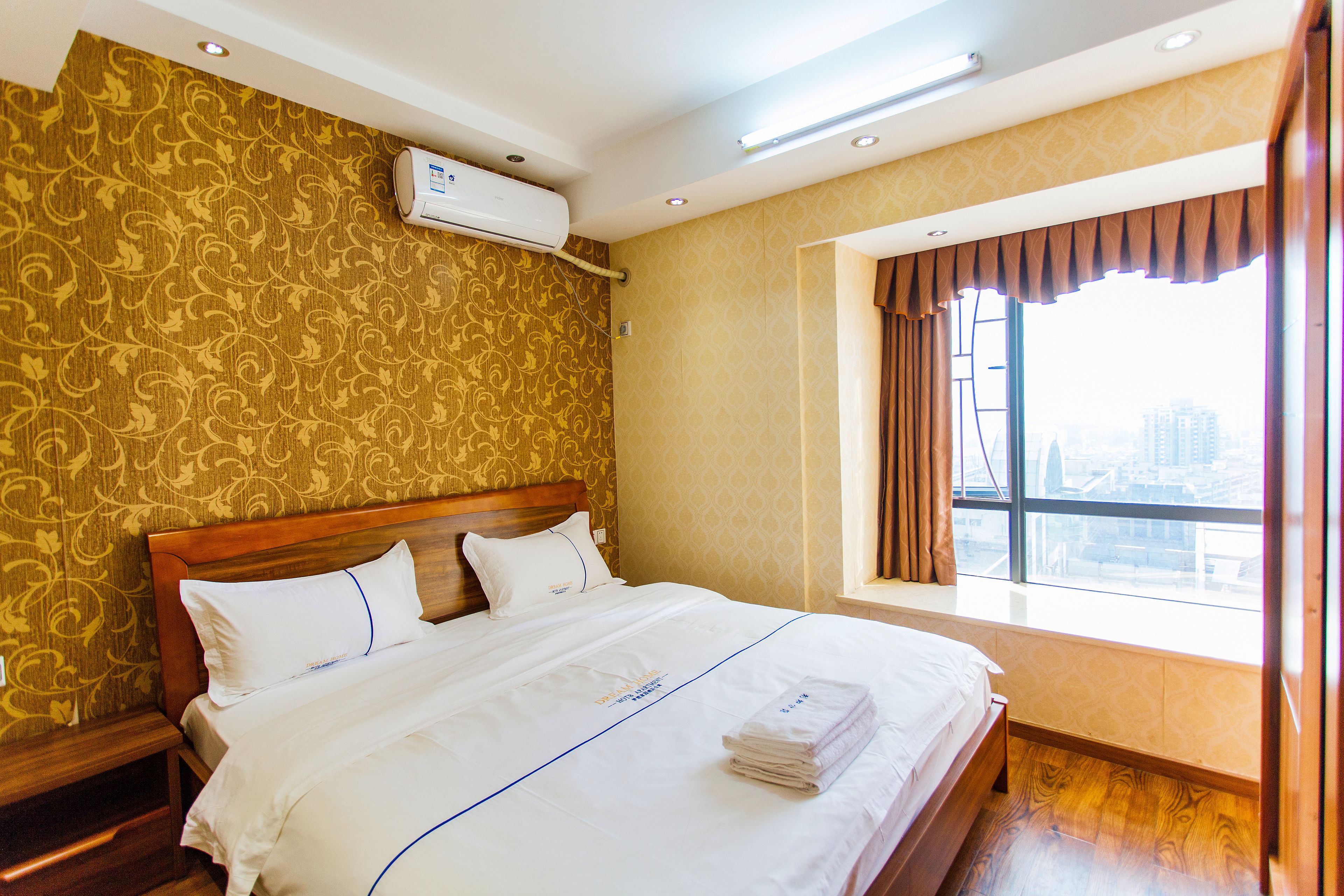 A hotel room with a large bed, a window with curtains, a wall-mounted air conditioner, and a patterned wallpaper.