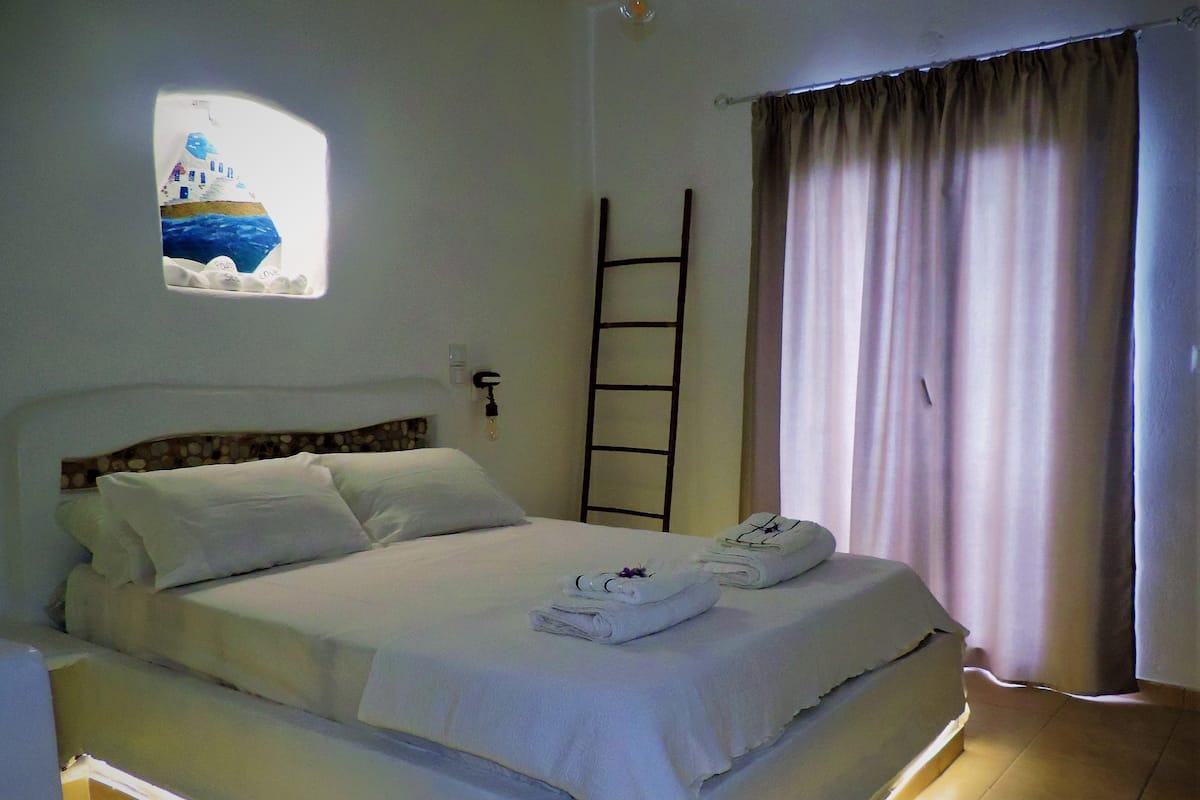 Una cama bien hecha con sábanas blancas, una ventana pequeña con un cuadro de una escena costera, una escalera apoyada en la pared y una cortina abierta a un lado.
