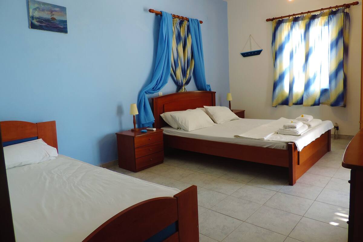 Habitación de hotel con dos camas de madera, una mesita de noche con lámpara y un escritorio, además de una ventana con cortinas.