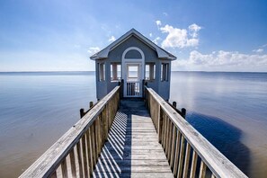 A wooden pier leading to a small, single-story building with a white door and a balcony, overlooking calm water.