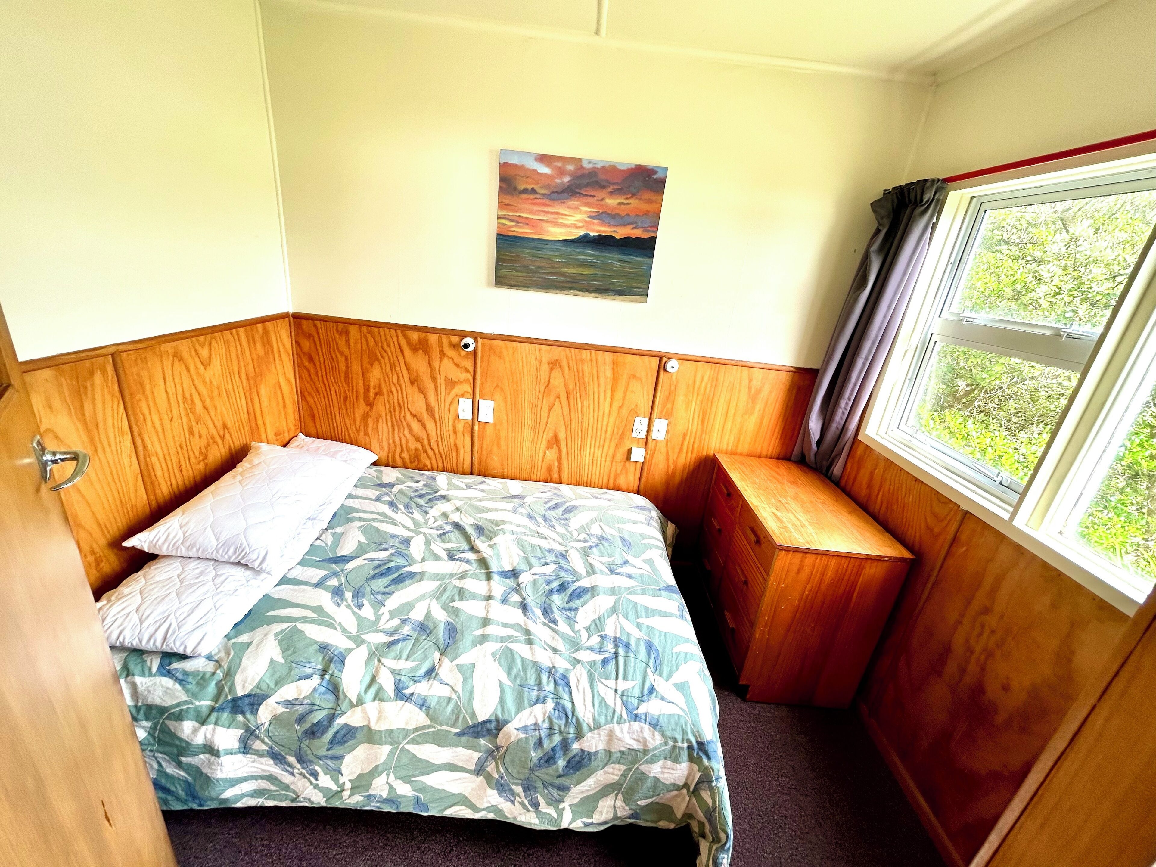 Una habitación pequeña con una cama, un banco de madera, una ventana con cortinas y un cuadro en la pared.