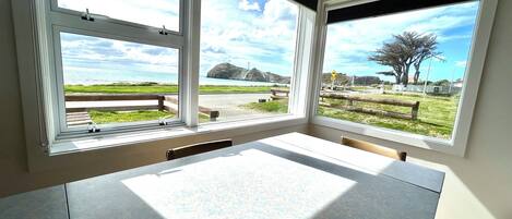 A room with a large table and three windows offering views of a coastal landscape with grassy areas and a road.