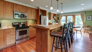 A kitchen with wooden cabinets, stainless steel appliances, a bar area with stools, and a view of the outdoors through large windows. - Charming Cottage in the Village of Warm Springs with Warm Thermal Spring (Warm Springs)