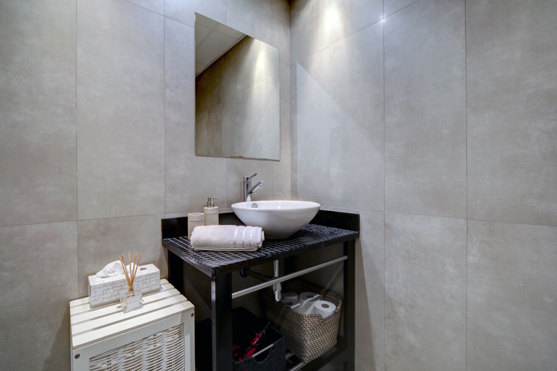 Une salle de bain moderne avec un grand lavabo blanc, un miroir et une étagère noire où sont rangés les articles de toilette.