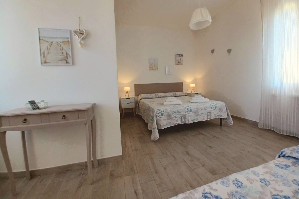 Quarto com cama, mesas de cabeceira, escrivaninha e luminária suspensa.
