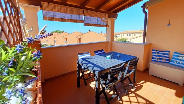 A covered patio with a table and chairs, a sofa, and a view of buildings in the background. - Il giardino (Piombino)