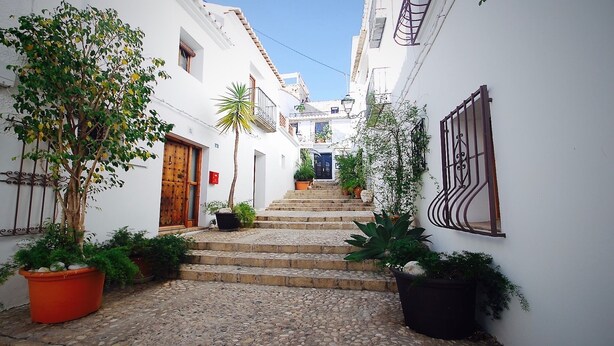 A narrow alleyway with white buildings, a wooden door, and steps leading to a blue door.
