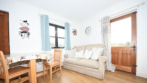 A cozy living room with a wooden floor, a beige sofa, a wooden dining table with chairs, and a window with blue curtains. - Self catering B&B or short holiday let (Saffron Walden)