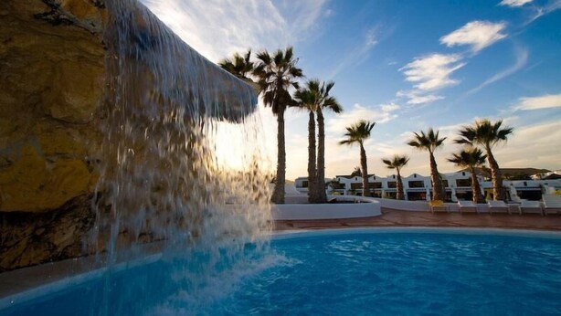 A pool with a waterfall feature, palm trees, and a clear sky. - Holiday Villa with shared pool in Costa Teguise, Las Cucharas (Costa Teguise)