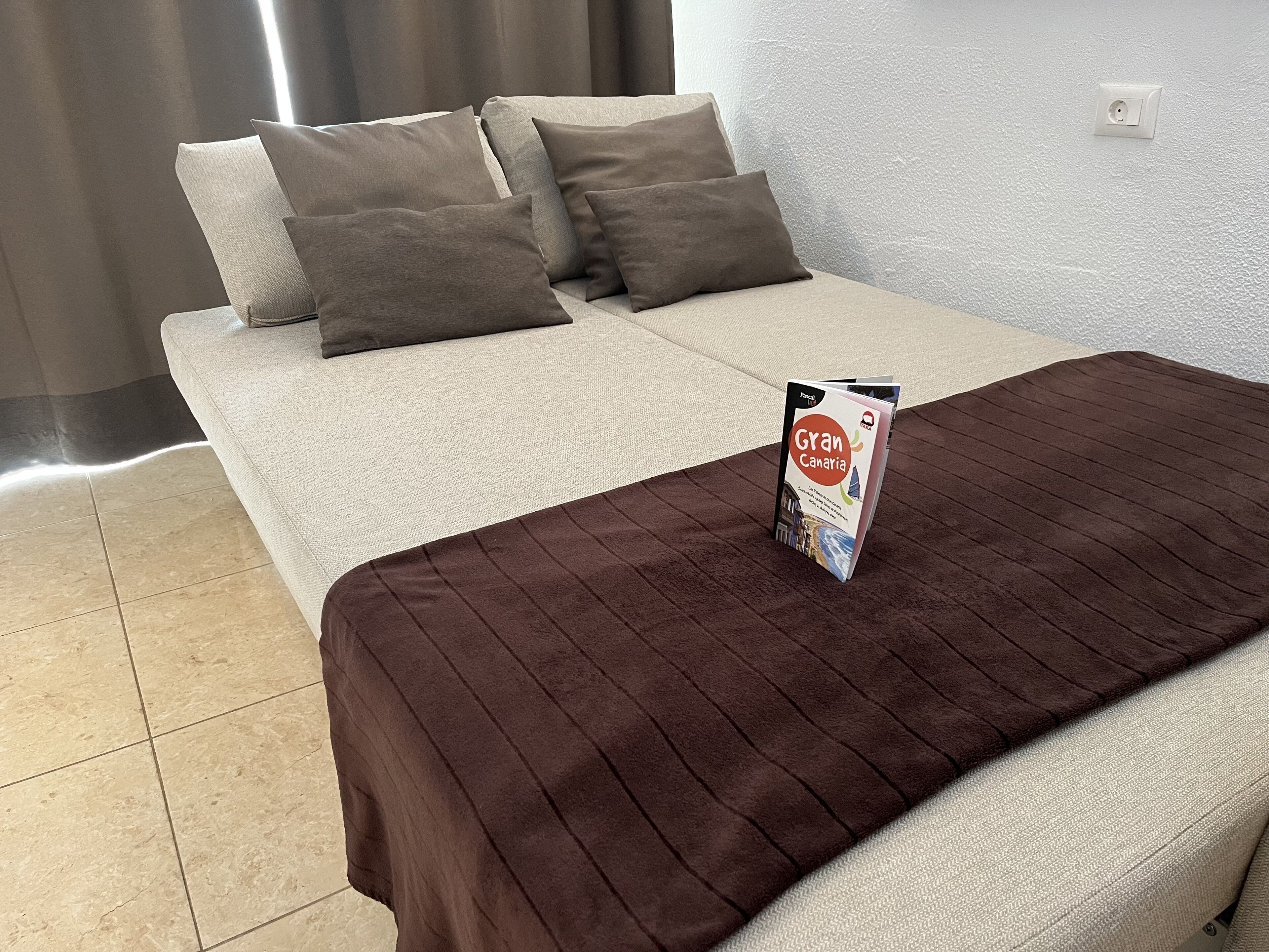 Ein Bett mit einer braunen Decke und einer Zeitschrift darauf. Im Hintergrund steht ein Sofa mit Kissen.