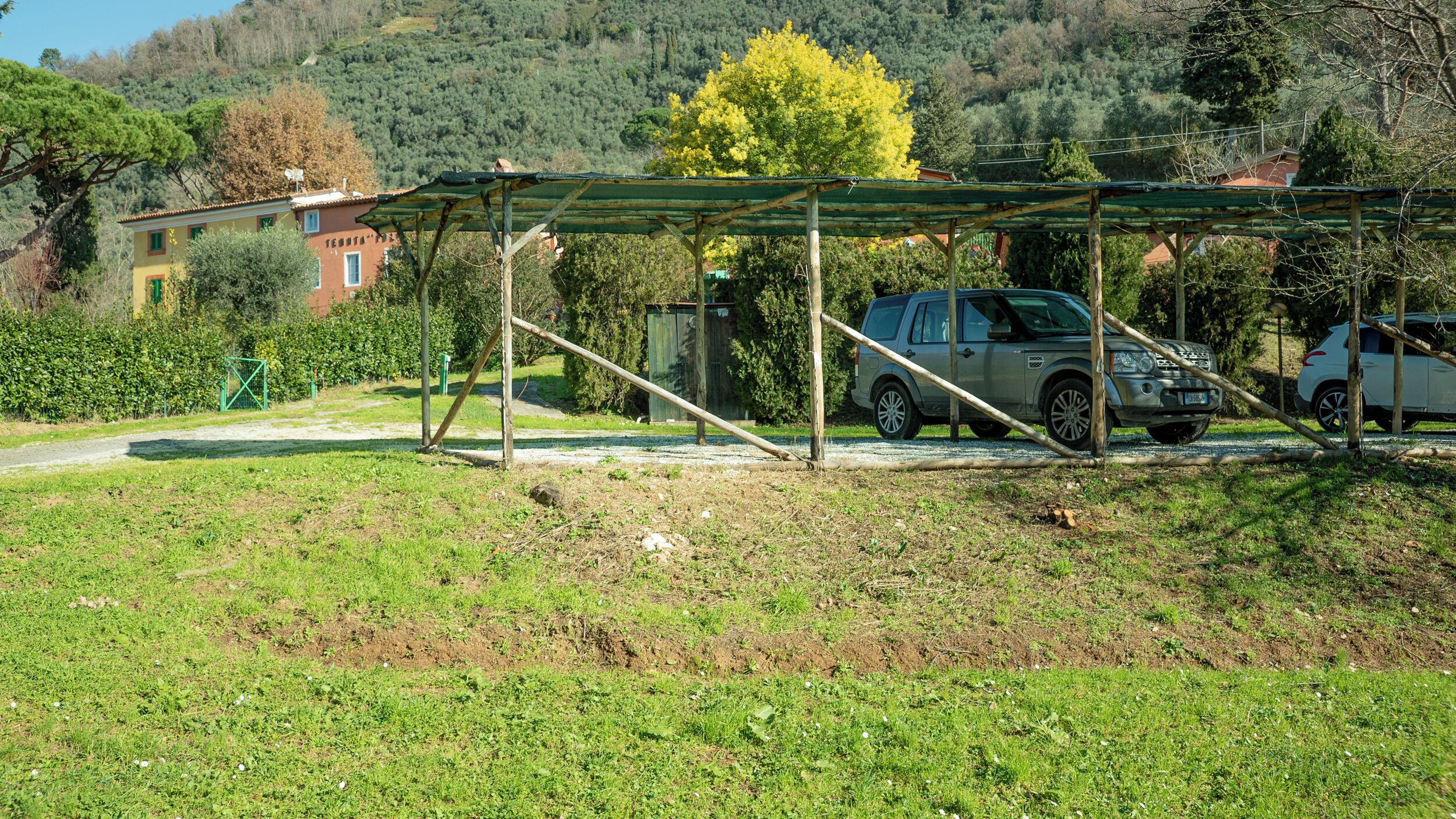 A parking area with a covered carport, a dirt path, and a building in the background.