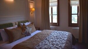 Un lit bien fait, recouvert d’une couvre-lit à motifs, avec une tête de lit verte et plusieurs oreillers. Il y a deux fenêtres avec des rideaux, et l’une d’elles offre une vue sur la verdure à l’extérieur.