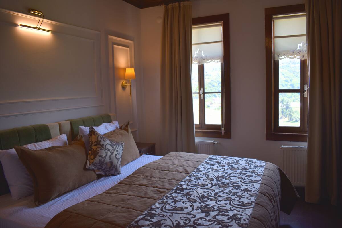 A neatly made bed with a patterned bedspread, a green headboard, and multiple pillows. There are two windows with curtains, one of which has a view of greenery outside.