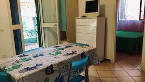 A room with a dining table covered with a floral tablecloth, a small white cabinet with drawers, a television on top, and a bed with a green bedspread.