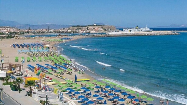 A beach with rows of sunbeds and umbrellas, a pier with a ship, and a cityscape in the background. - Charming Venetian house in heart of Rethymno (Rethymno)
