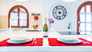 Une table à manger est dressée avec des assiettes, des couverts et une bouteille de champagne. Un set de table rouge est placé sous chaque assiette. La pièce comprend une cuisine moderne avec une pancarte « Cuisine » sur le mur, une grande horloge et une fenêtre avec des rideaux.