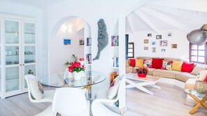 Un vaste espace de vie comprenant une table à manger en verre, des chaises blanches, un canapé agrémenté de coussins colorés et une bibliothèque.