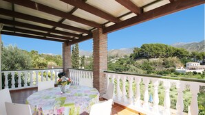 Eine überdachte Terrasse mit Tisch und Stühlen, einem weißen Geländer und Blick auf eine grüne Landschaft mit Bäumen und Hügeln.