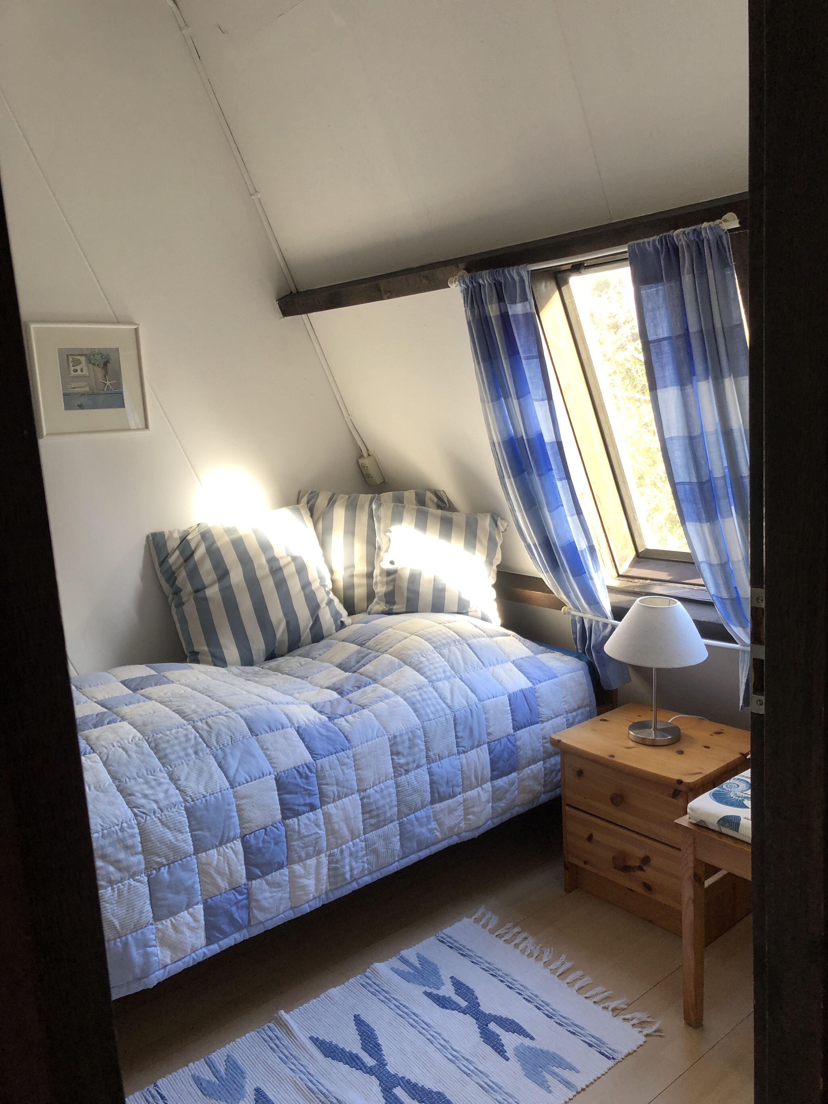 Quarto com cama, mesa de cabeceira com luminária, pequena gaveta e janela com cortinas.