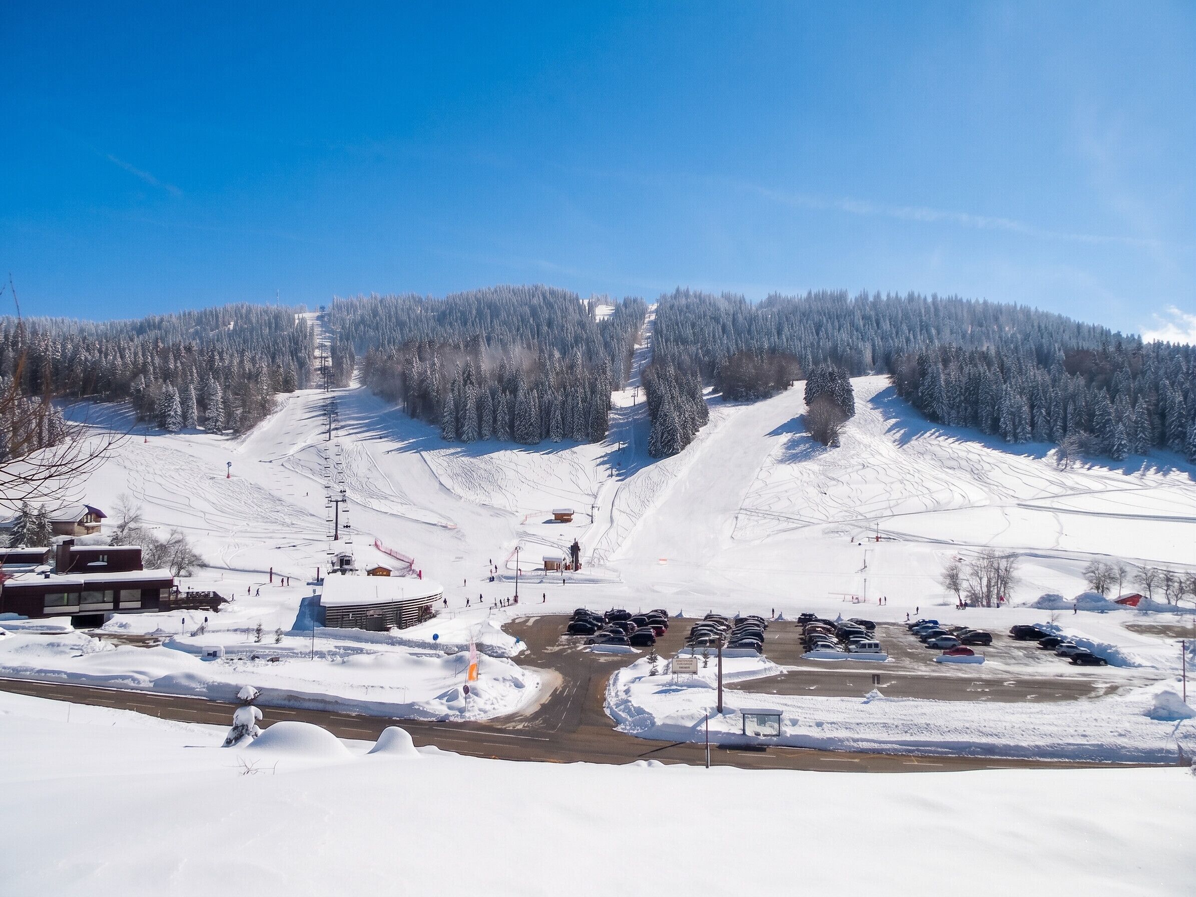 Estación de esquí cubierta de nieve con zona de estacionamiento, remontes y el paisaje de una montaña.